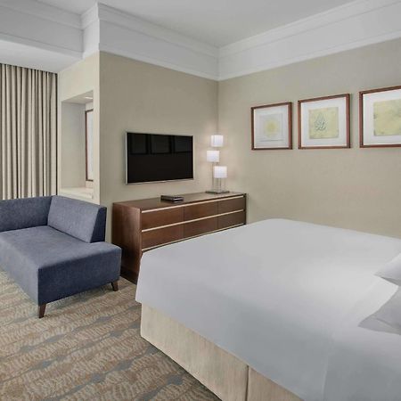 Hotel Jabal Omar Hyatt Regency Makkah La La Mecca Esterno foto The photo shows a modern hotel room featuring a neatly made bed with white linens, a small blue couch to the side, and a wooden dresser with a television mounted on the wall above it. There are two framed pieces of art on the wall near the bed, and t