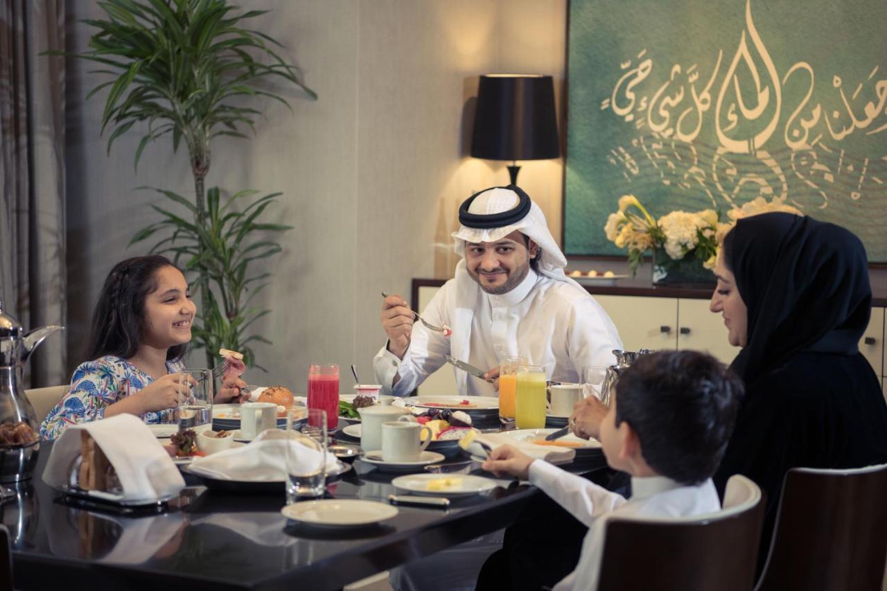 Hotel Jabal Omar Hyatt Regency Makkah La La Mecca Esterno foto The photo depicts a cheerful family gathered around a dining table for a meal. They appear to be enjoying breakfast or a brunch. The table is set with various dishes and drinks, including juices and plates of food. 

A man wearing traditional attire 