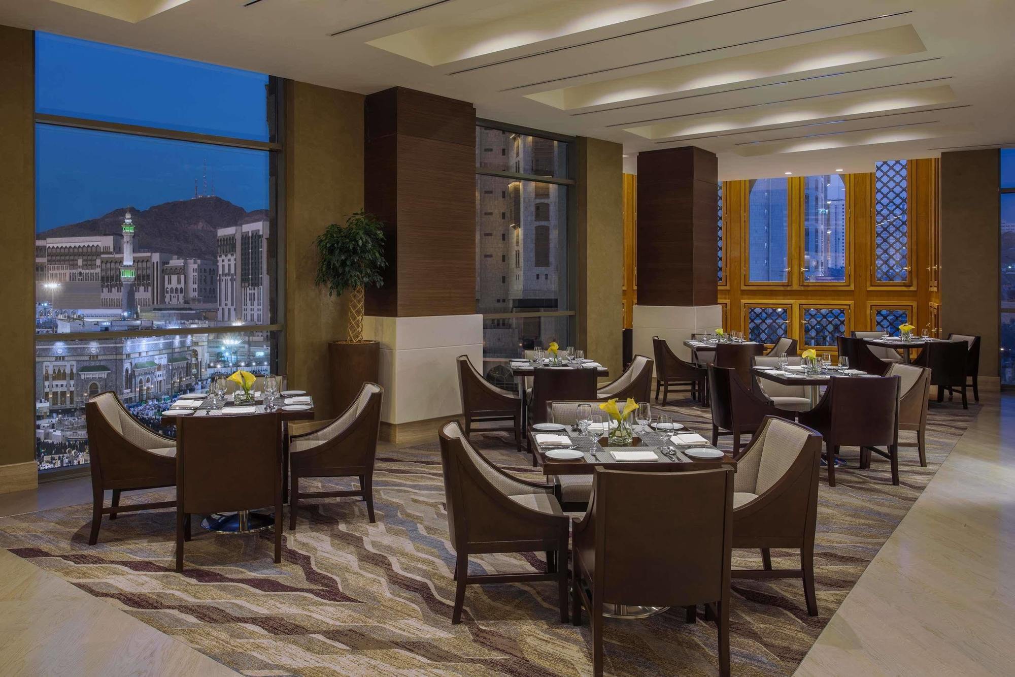 Hotel Jabal Omar Hyatt Regency Makkah La La Mecca Esterno foto The photo shows a sophisticated restaurant interior, featuring elegant round tables with high-backed chairs arranged neatly. The space is well-lit with large windows that provide a view of the city skyline at dusk, with mountains in the background. T
