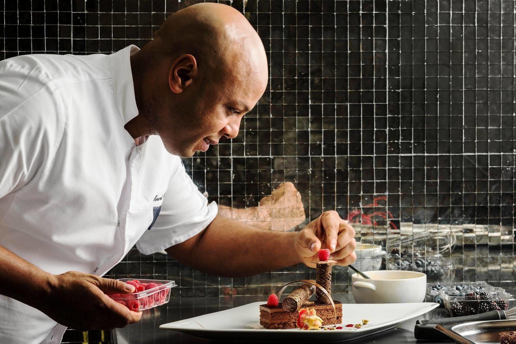 Hotel Jabal Omar Hyatt Regency Makkah La La Mecca Esterno foto The photo depicts a chef, who is bald and wearing a white chef's coat, carefully plating a dessert. He is focused on adding a finishing touch, possibly a raspberry, to the dessert on a large white plate. The background features a tiled wall, and ther