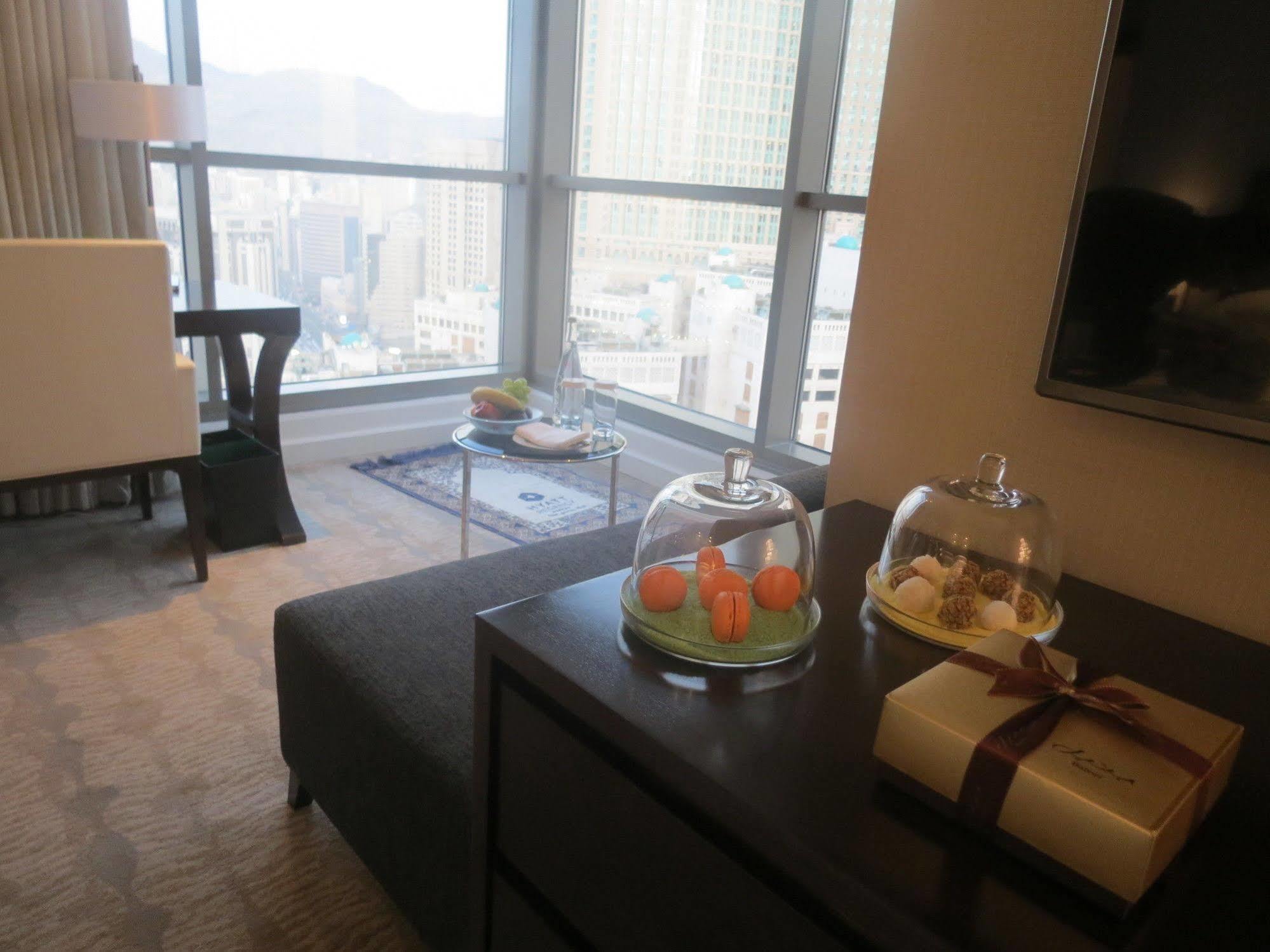 Hotel Jabal Omar Hyatt Regency Makkah La La Mecca Esterno foto The photo shows a modern hotel room or a stylish living space with large windows offering a view of the city. In the foreground, there is a dark sofa and a low table with covered trays displaying what appears to be small desserts or delicacies. To th
