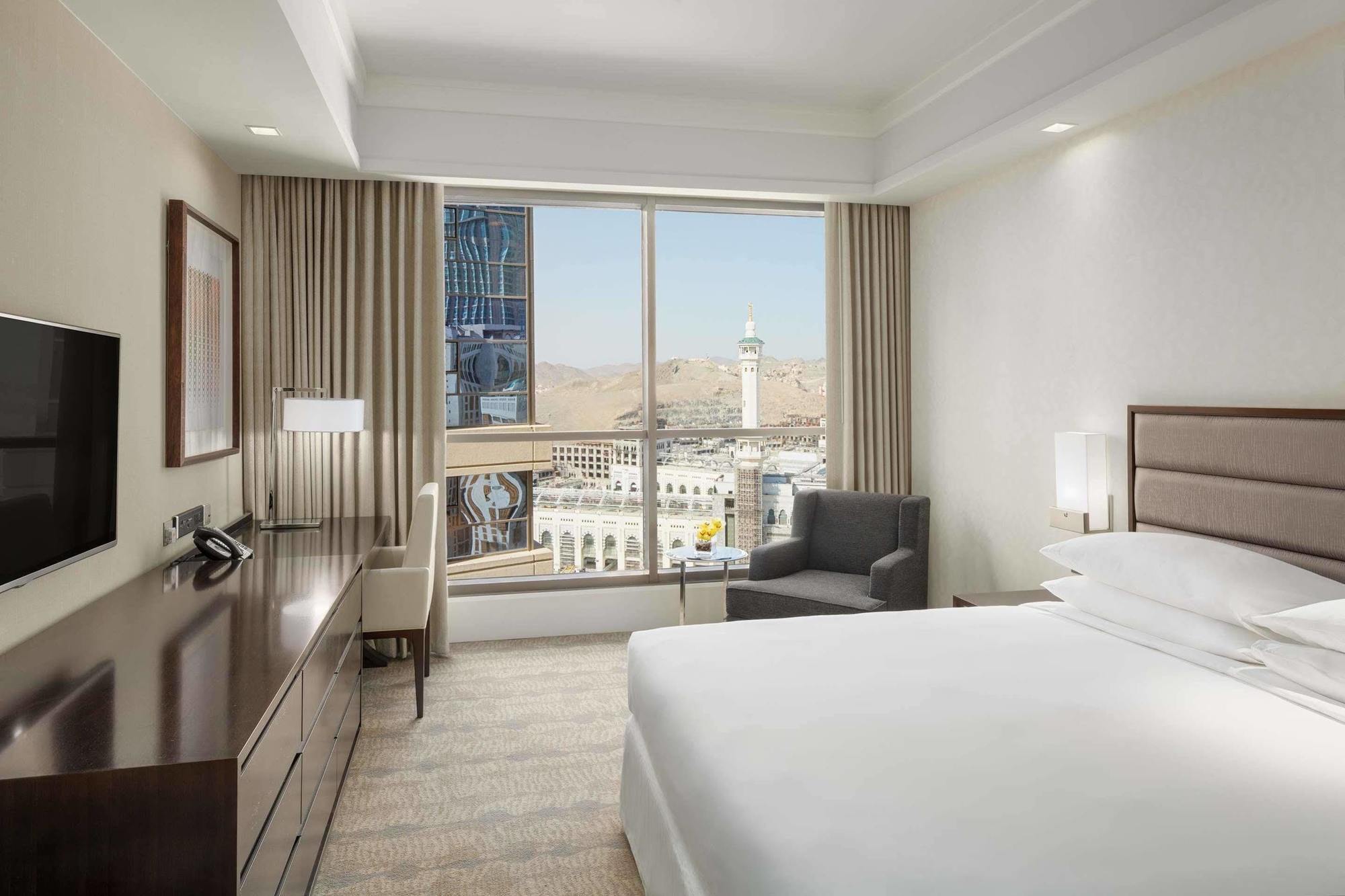 Hotel Jabal Omar Hyatt Regency Makkah La La Mecca Esterno foto The photo shows a modern hotel room featuring a large window that provides a view of an outdoor landscape, likely part of a city or landmark. The room includes a bed with white linens, a side table with a lamp, and a small seating area with a chair a