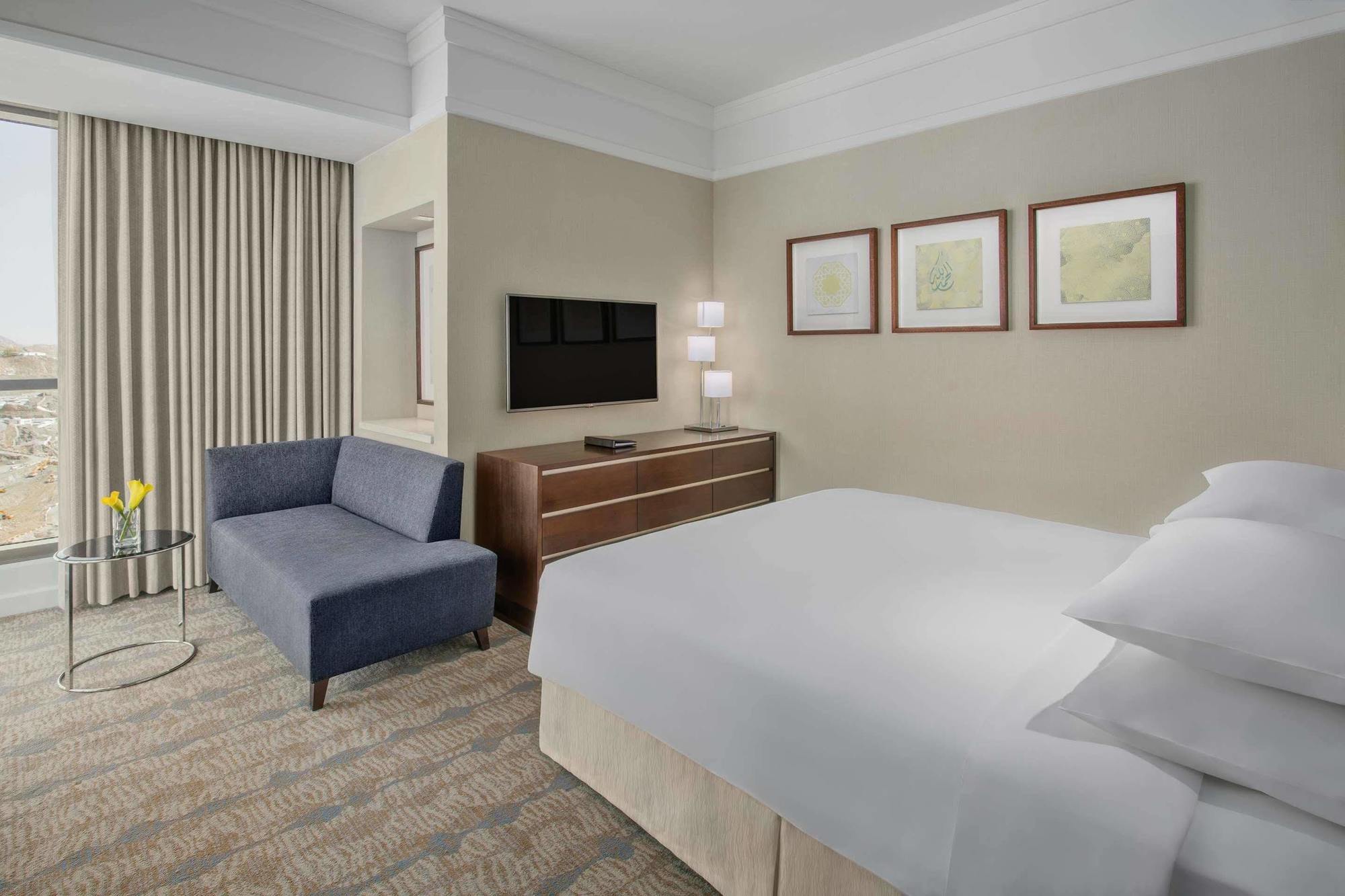 Hotel Jabal Omar Hyatt Regency Makkah La La Mecca Esterno foto The photo shows a modern hotel room featuring a neatly made bed with white linens, a small blue couch to the side, and a wooden dresser with a television mounted on the wall above it. There are two framed pieces of art on the wall near the bed, and t