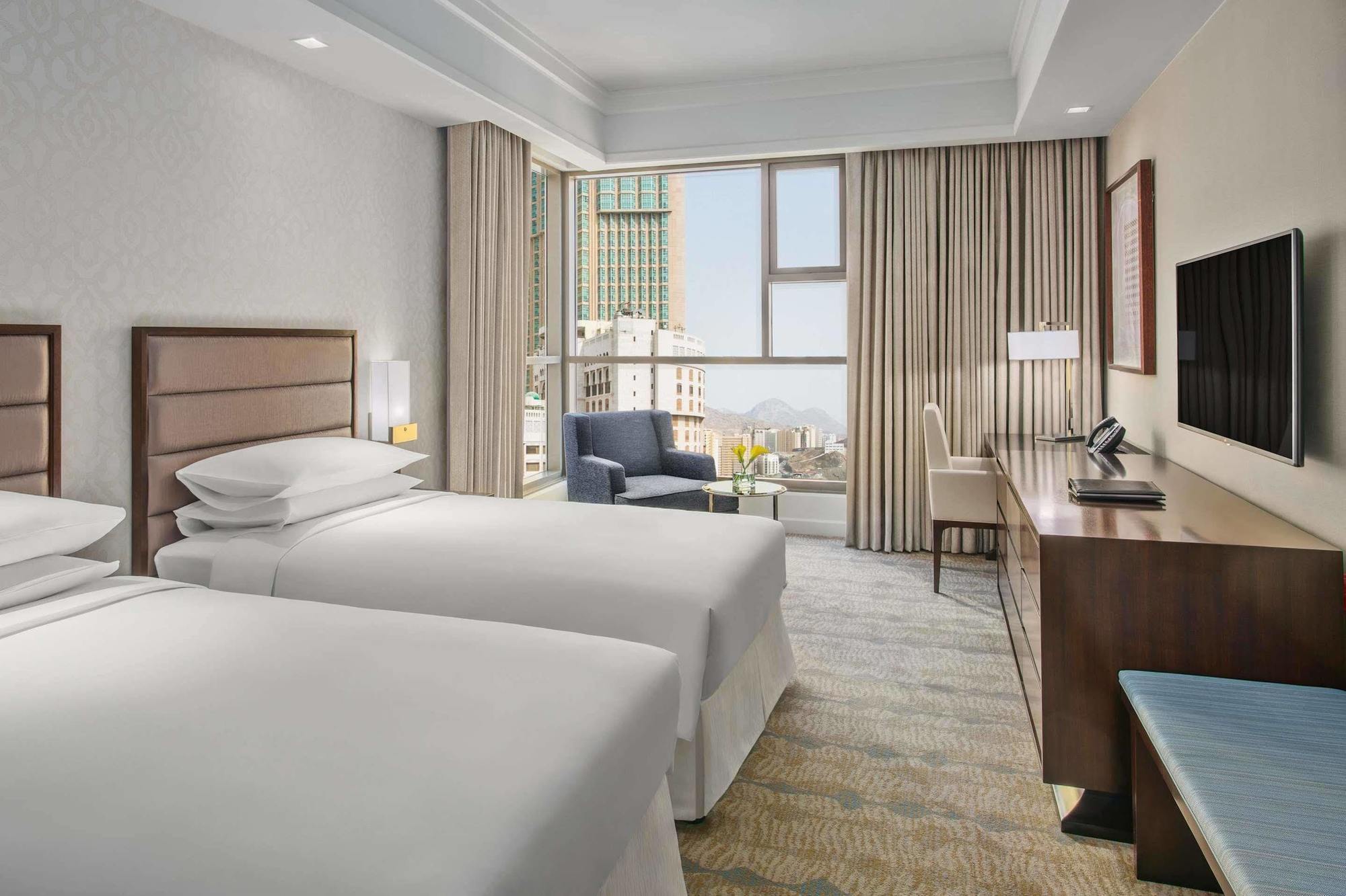 Hotel Jabal Omar Hyatt Regency Makkah La La Mecca Esterno foto The photo shows a modern hotel room featuring two single beds with white bedding. There's a spacious window allowing natural light to fill the room, which is adorned with light-colored curtains. In one corner, there's a comfortable armchair next to a