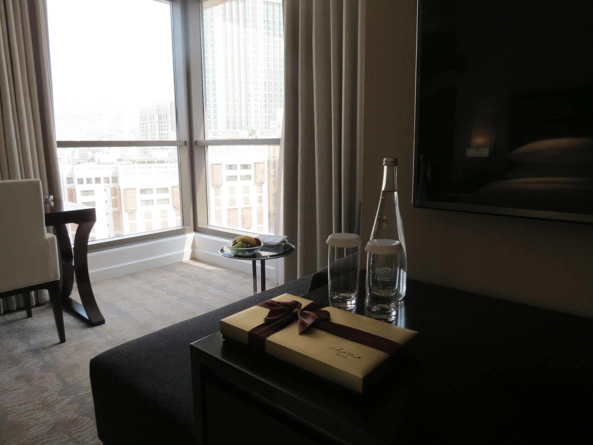 Hotel Jabal Omar Hyatt Regency Makkah La La Mecca Esterno foto The photo shows a modern hotel room with large windows letting in natural light. In the foreground, there is a table with a decorative box tied with a ribbon, two clear glass water bottles, and a couple of glasses. In the background, there's a comfor